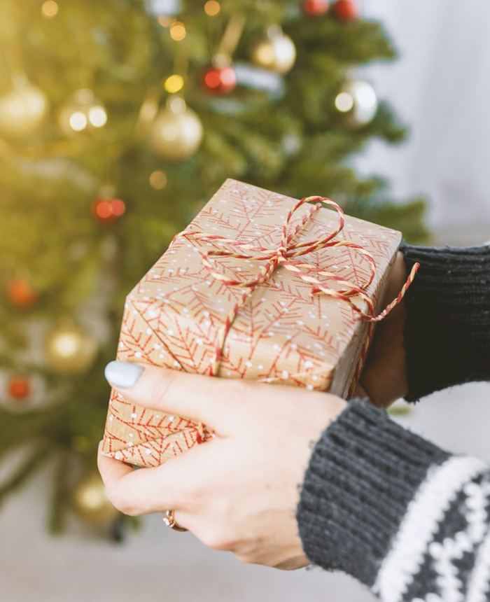 Hands holding a kraft-paper wrapped gift beside a Christmas tree